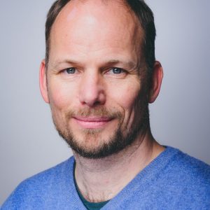 Portraitaufnahme von Philipp Wetzel. Er trägt einen hellblauen Pullover und lächelt in die Kamera.